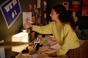 Uma jovem estudante organiza seu cronograma de estudos em um quadro fixado na parede do quarto, escrevendo com caneta colorida enquanto sorri. A mesa está iluminada e repleta de materiais escolares, como cadernos, canetas e fones de ouvido.