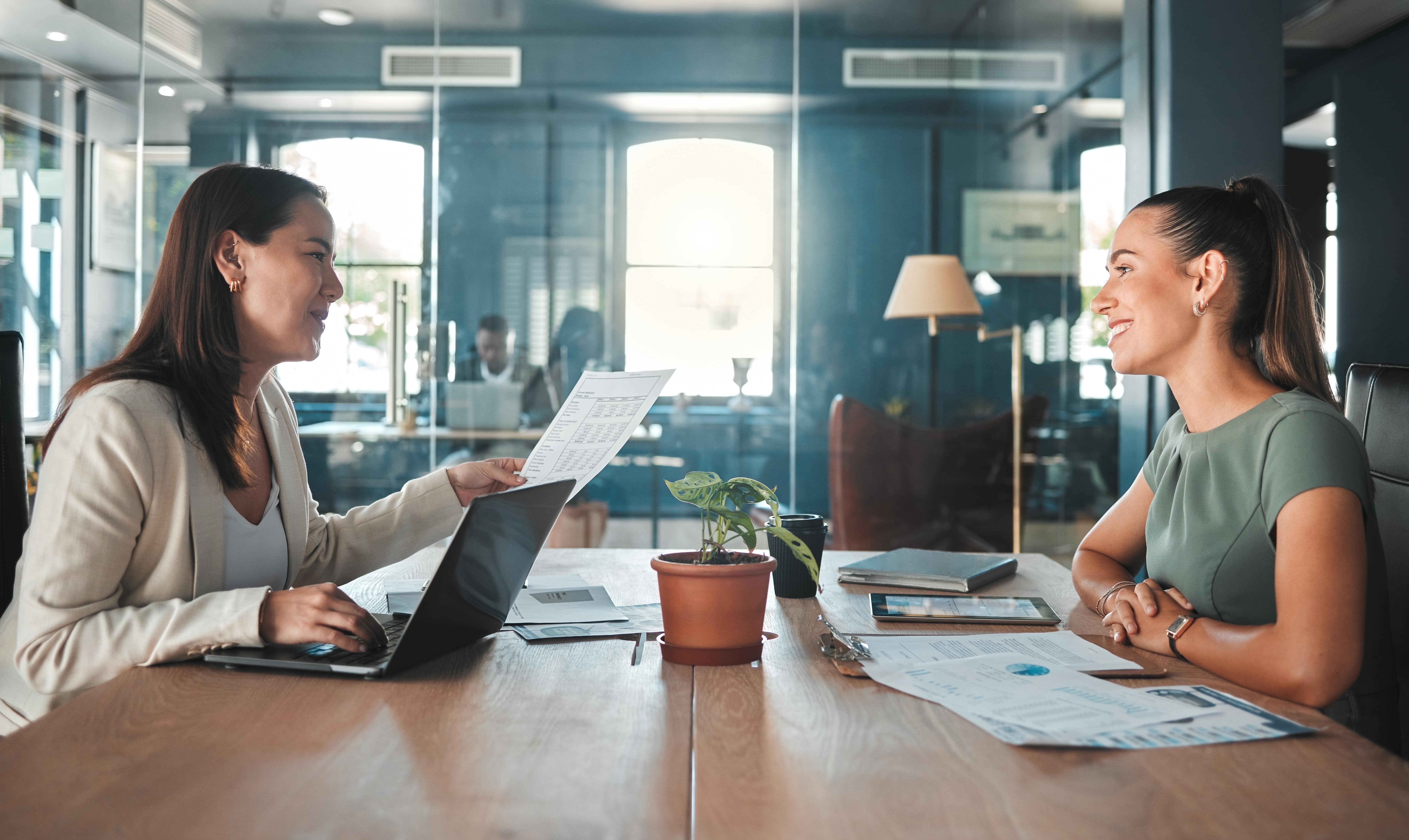 Mulher jovem sendo entrevistada em ambiente corporativo moderno, sentada à mesa com uma recrutadora que analisa seu currículo. A cena transmite profissionalismo, preparação e confiança — ideal para ilustrar conteúdos sobre como fazer um bom currículo.