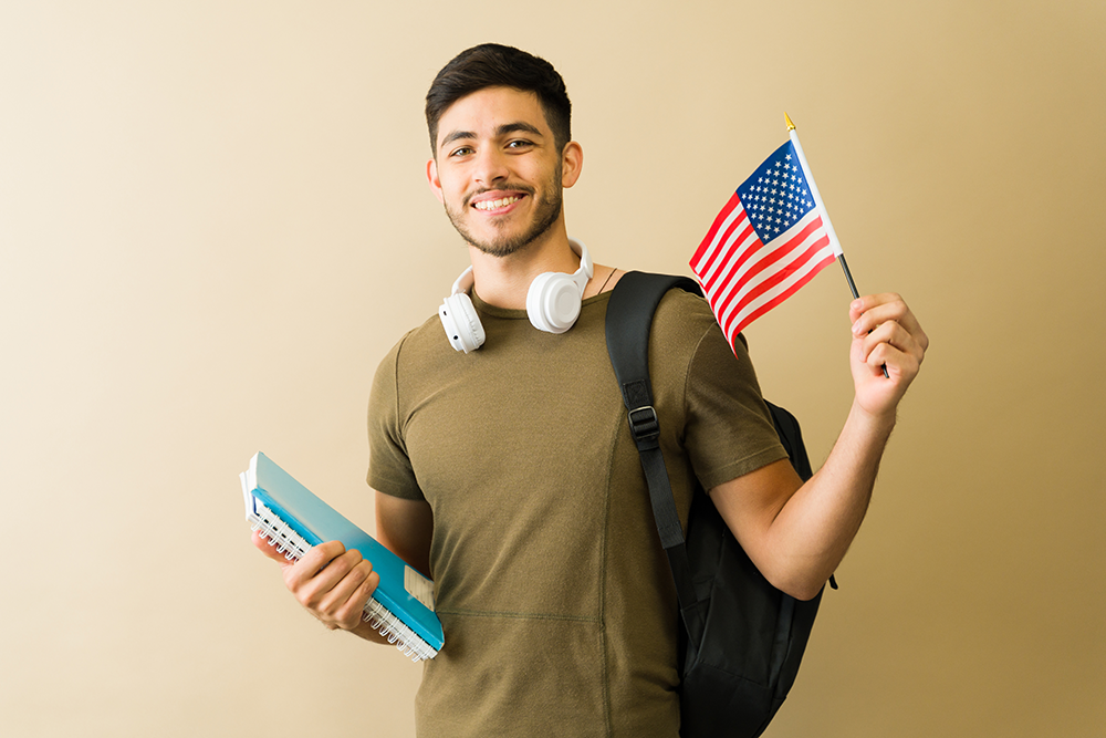 Jovem estudante sorridente com mochila e cadernos. Ele usa fones de ouvido e segura a bandeira dos EUA. Isso representa motivação e foco. O objetivo é descobrir suas habilidades profissionais através do inglês. A imagem transmite determinação e preparo pa