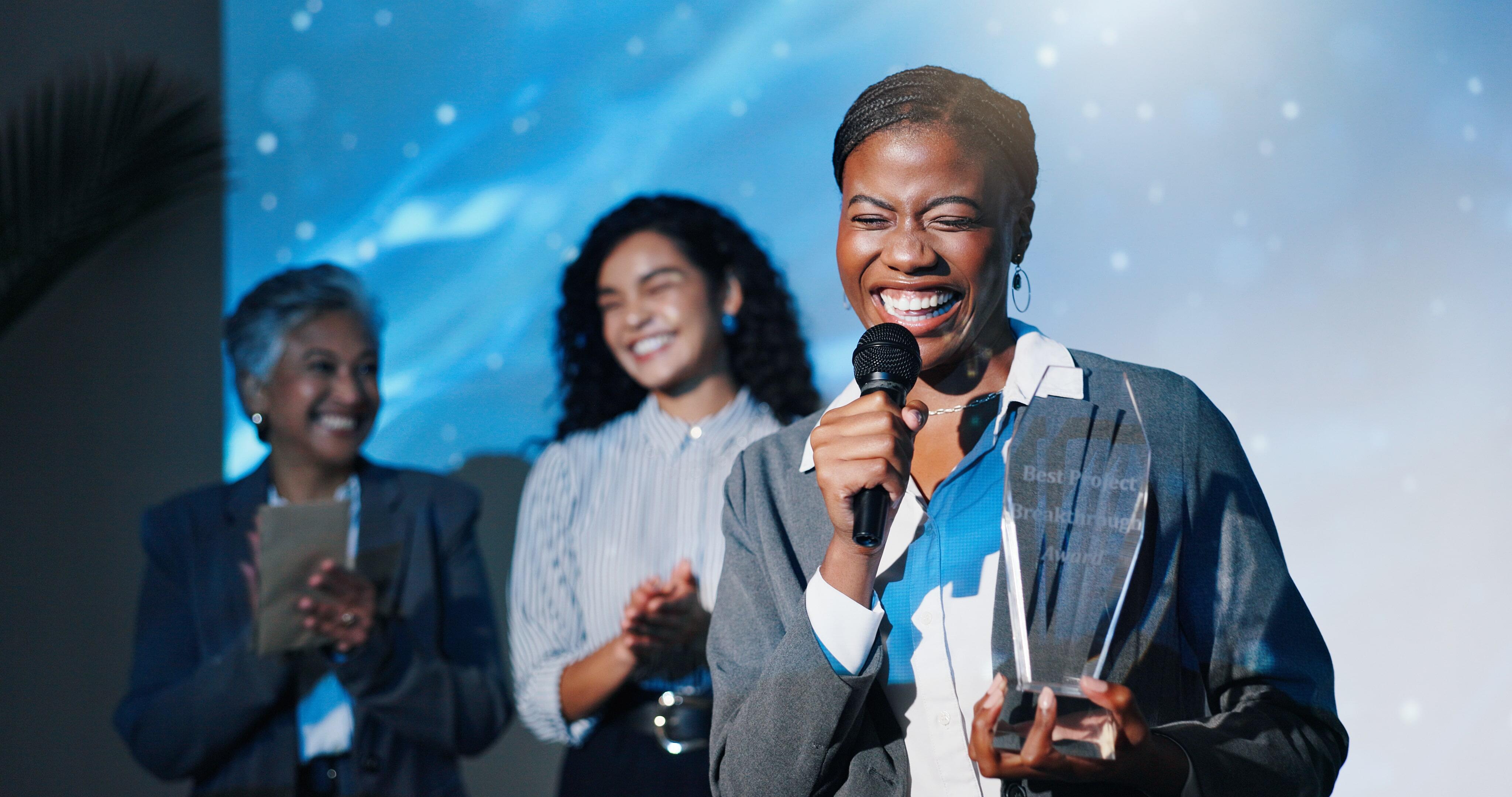 Mulher negra sorrindo enquanto fala ao microfone em uma cerimônia de premiação, segurando um troféu. Ao fundo, duas colegas aplaudem com entusiasmo. A imagem transmite superação, autoconfiança e destaque profissional.