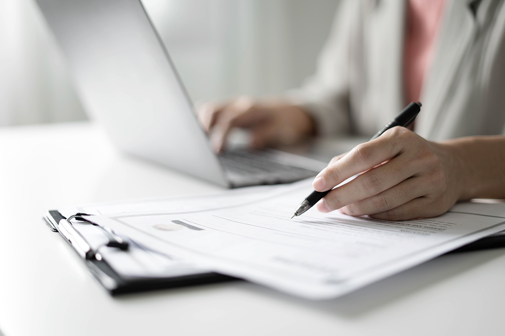 imagem de uma pessoa com uma mão no teclado de um notebook no fundo e a outra mão em destaque com uma caneta em cima de alguns papéis
