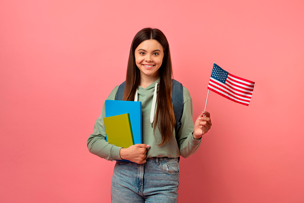 Uma jovem sorridente segura uma bandeira dos Estados Unidos e cadernos escolares, representando estudantes que desejam aprender inglês. A imagem ilustra bem o tema "o que impede de aprender inglês" , destacando a motivação e os recursos como fatores impor