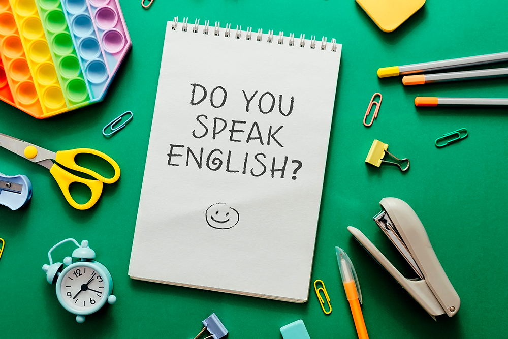 Fundo verde com um caderno em cima escrito "Do you speak inglish?" (você fala inglês). Em volta vários materiais de estudo como lápis, borracha, tesoura e outros.
