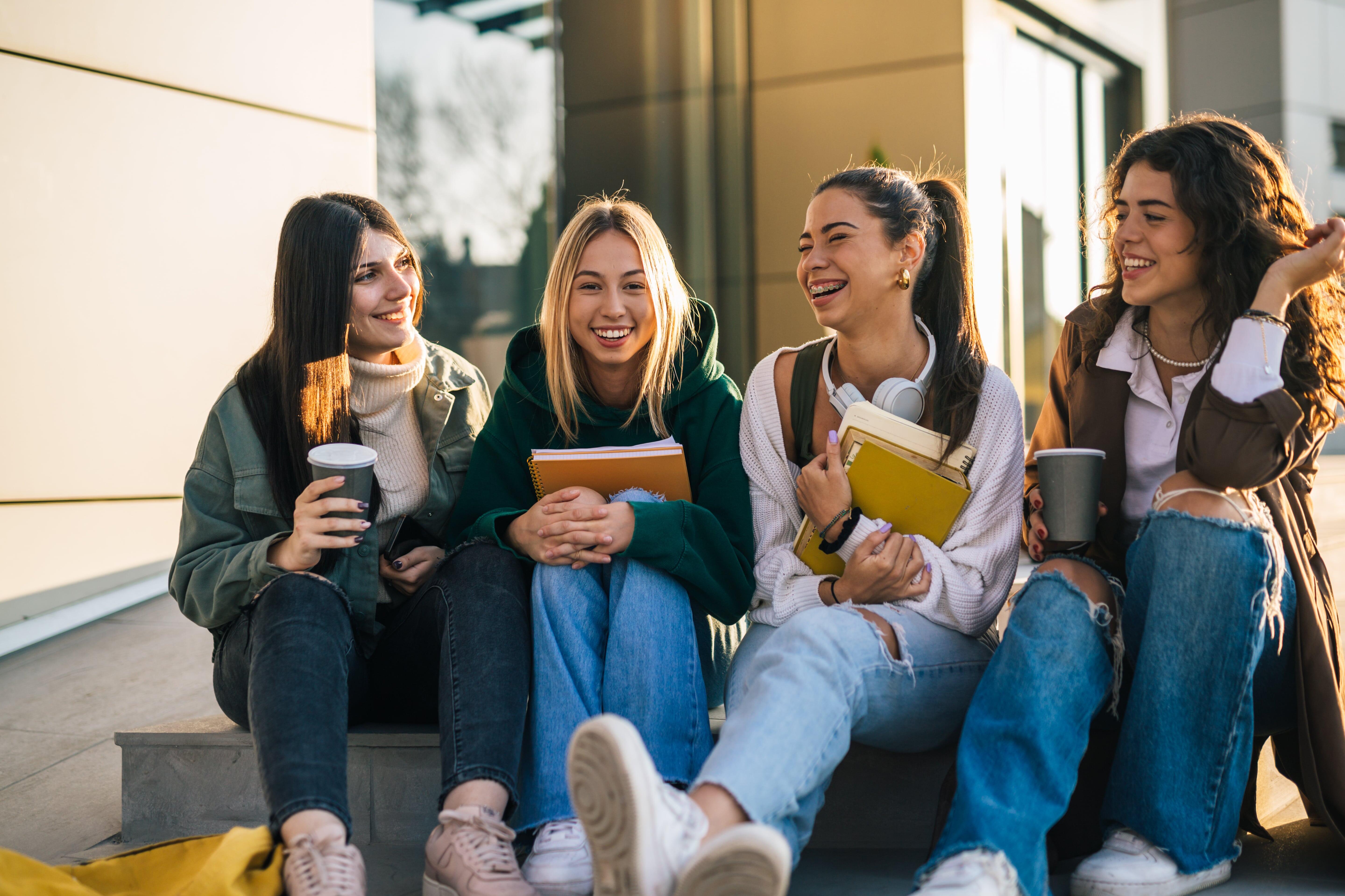 Grupo de jovens sorrindo sentadas na calçada com cadernos e café nas mãos, representando estudantes que podem se beneficiar de aprender expressões em inglês para destacar seus currículos.