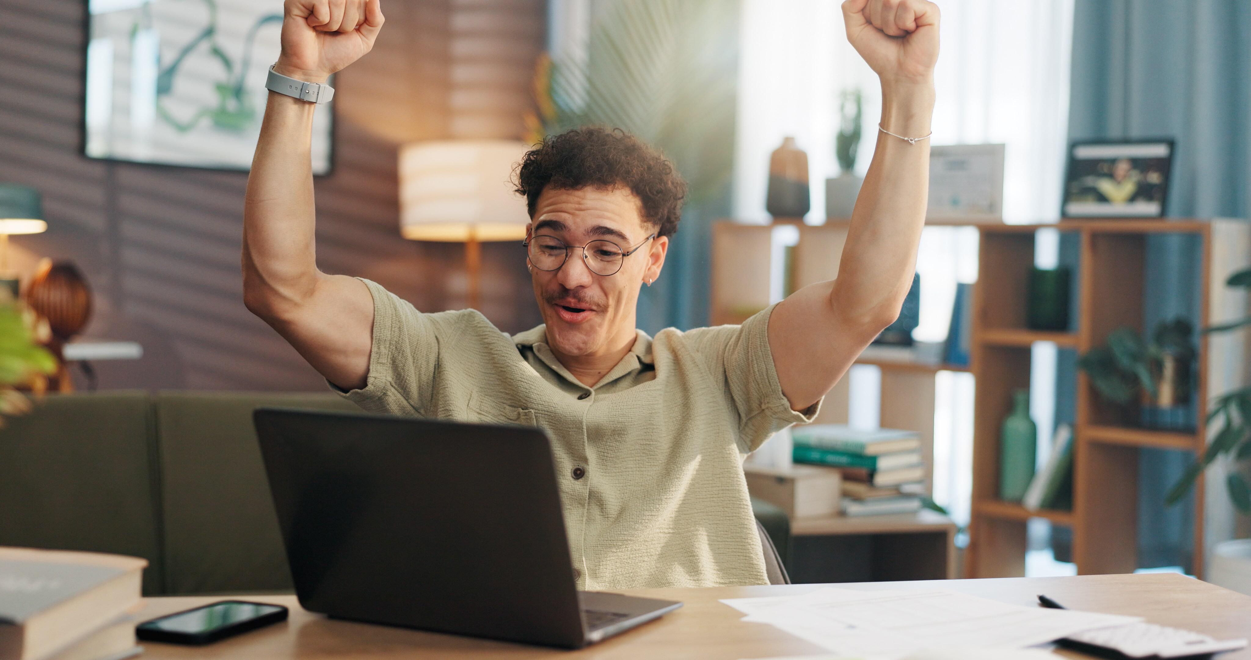 Homem jovem de óculos e bigode comemorando em frente a um computador, ele parece estar em home office.
