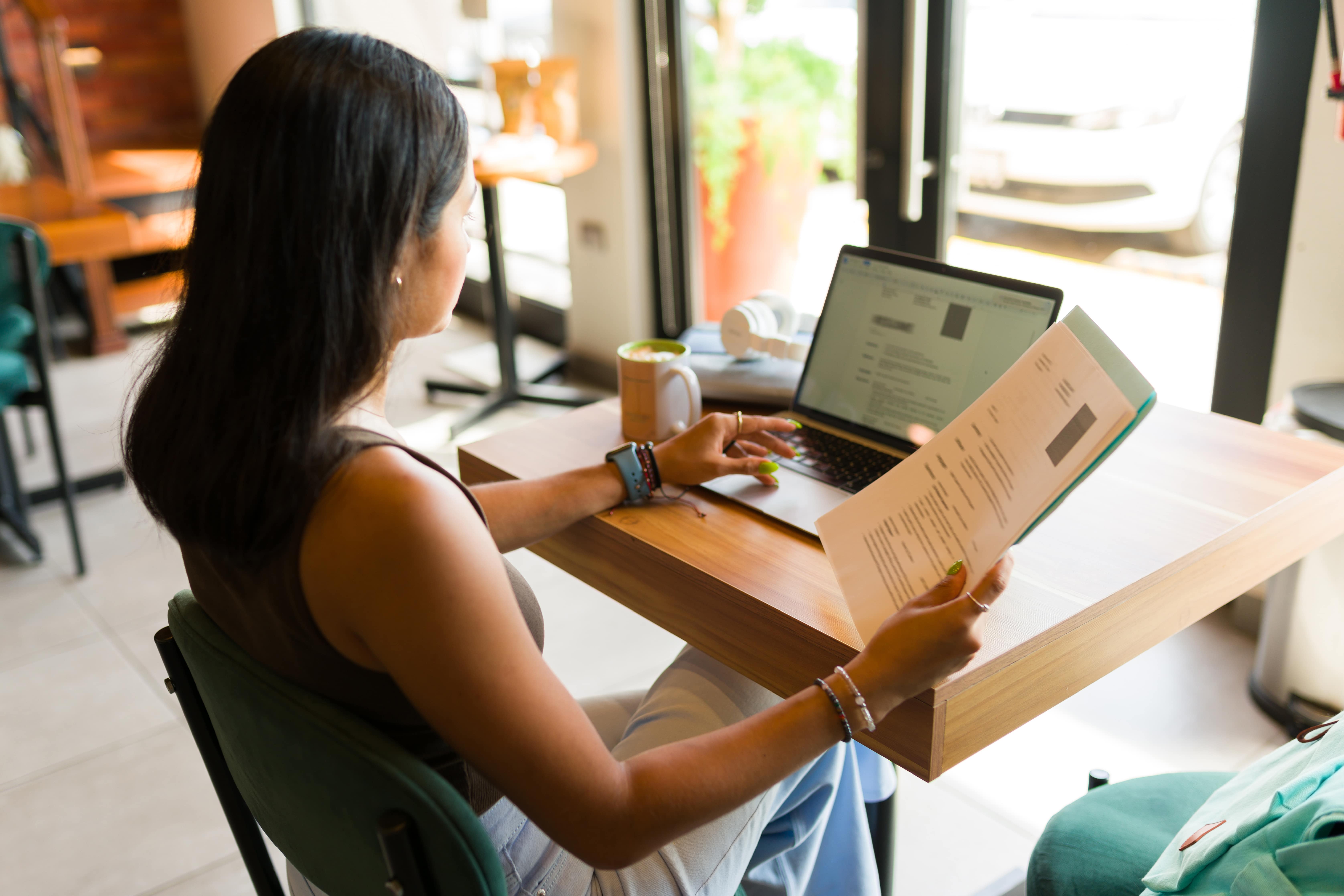Jovem ajustando o currículo no notebook enquanto segura uma versão impressa, simbolizando o cuidado ao escrever o objetivo profissional.
