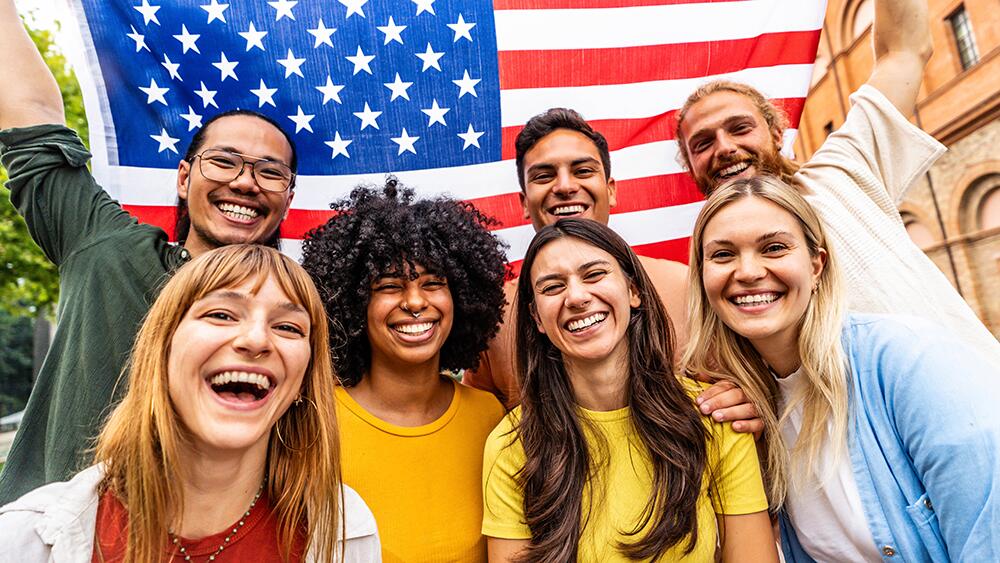 Grupo de jovens sorridentes de diferentes origens, reunidos em frente a uma bandeira dos Estados Unidos, representando diversidade e conexão com o idioma inglês. A imagem reforça a importância do inglês para o mercado de trabalho, especialmente em context