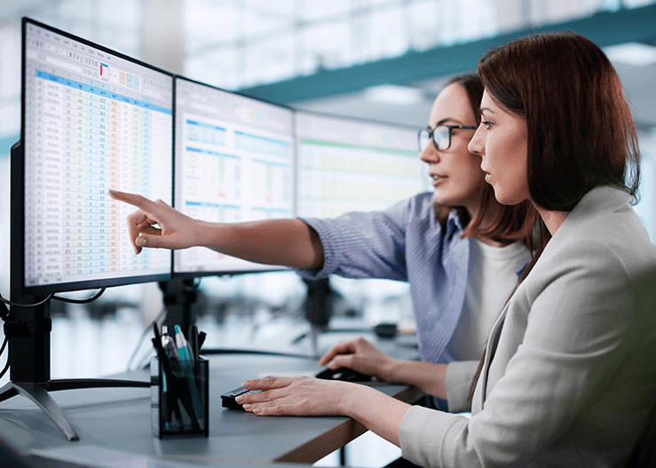 A imagem mostra duas profissionais analisando planilhas em monitores no escritório, representando o ambiente de trabalho típico ao discutir quanto ganha um programador