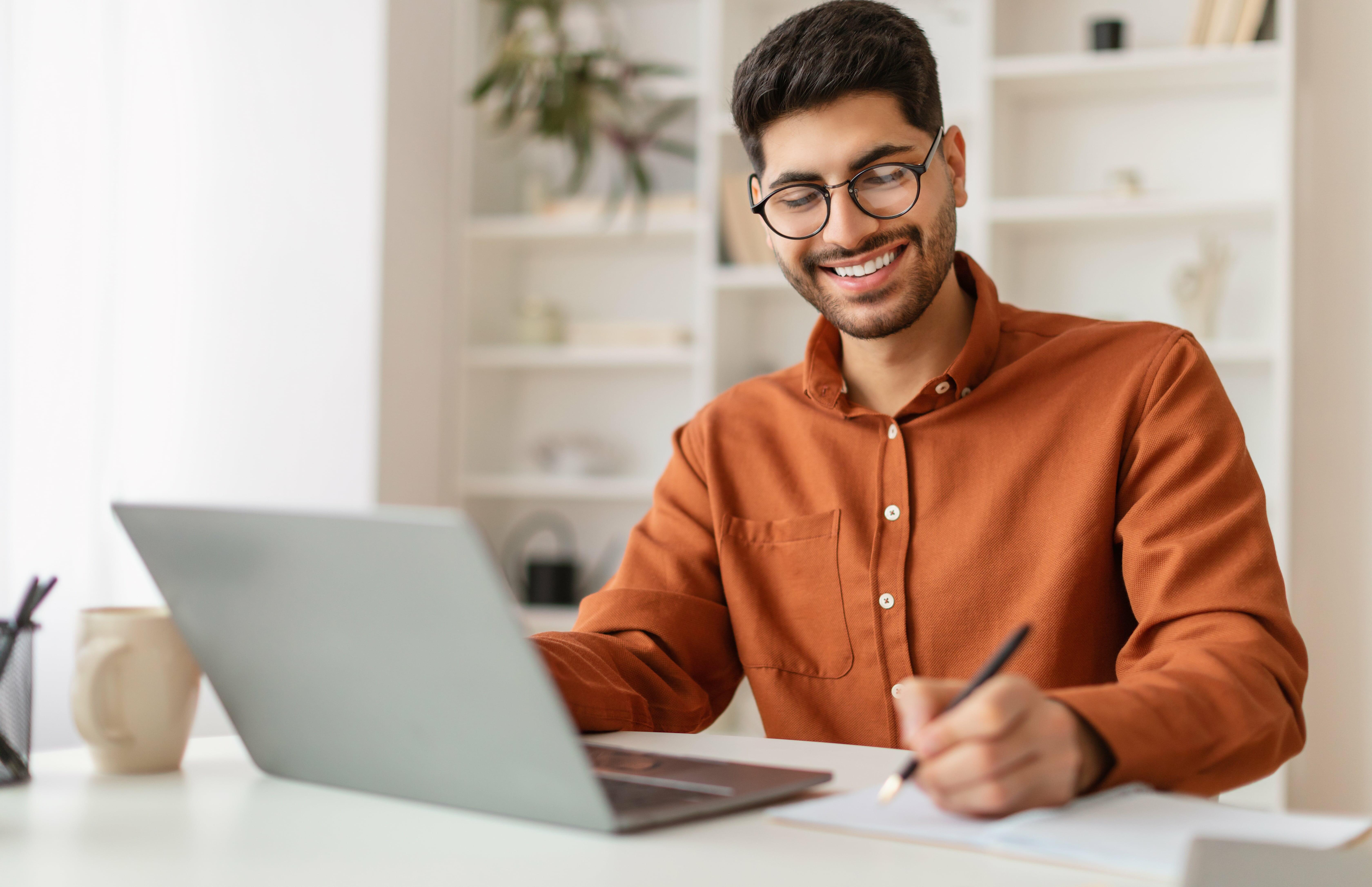 Jovem sorridente estudando em casa no computador e fazendo anotações, representando os 4 passos para se conhecer melhor no dia a dia e a importância do autoconhecimento na vida profissional